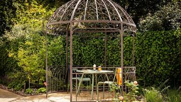 Un kiosque en métal surmonté d’une toiture en verre, entouré d’une végétation luxuriante et bordé d’un chemin de gravier.