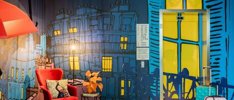 A room with a mural of a cityscape, a red armchair, a small table with a plant, and a suitcase.