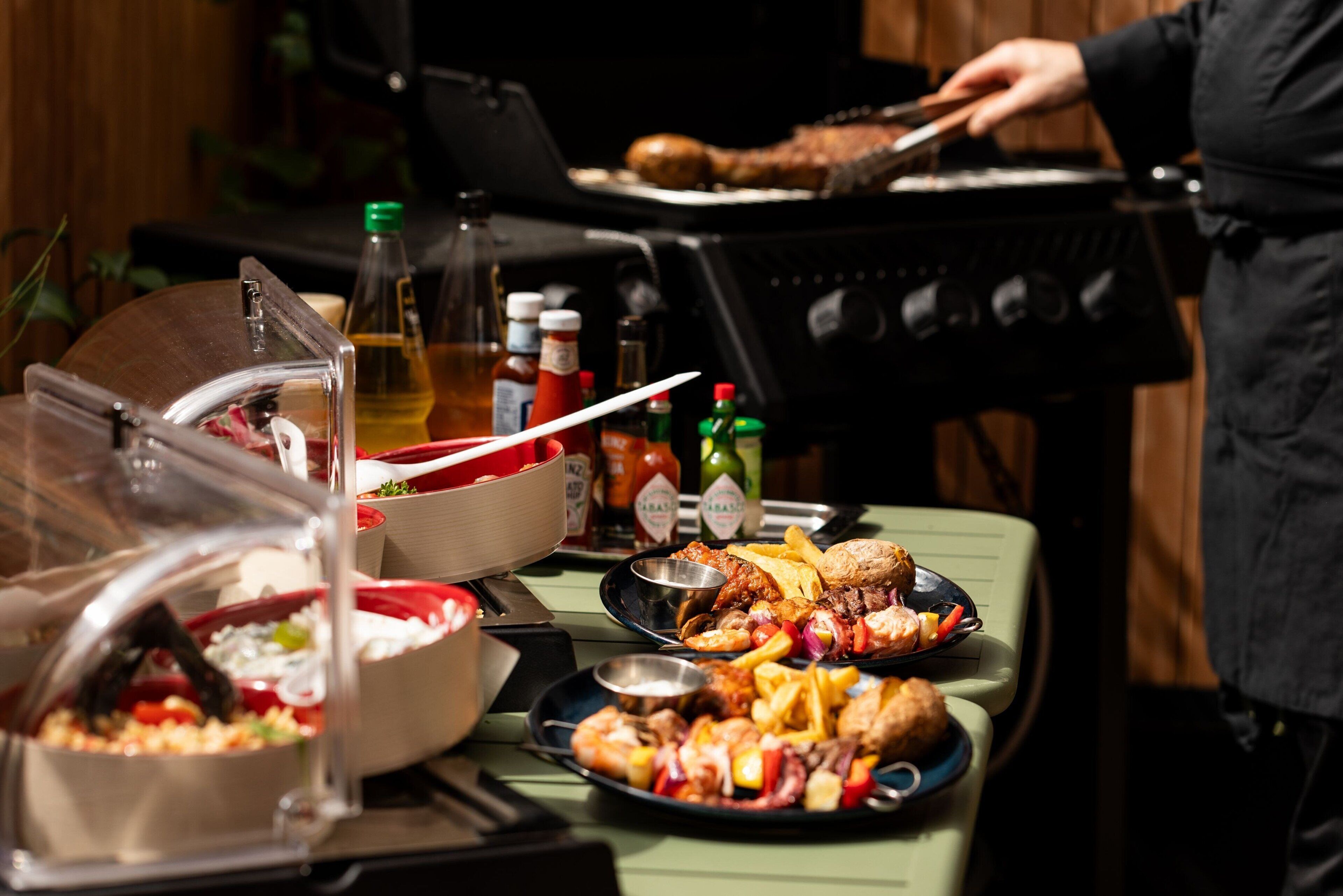 A buffet table with various dishes, a grill in the background, and condiments.
