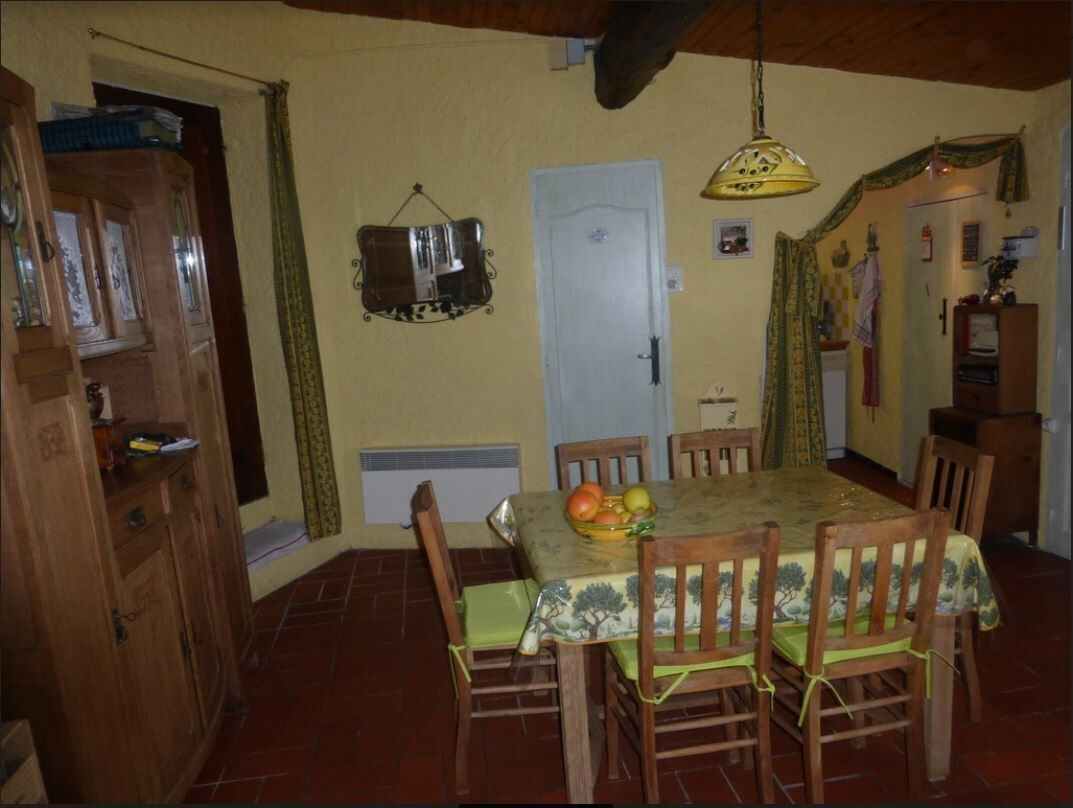 A dining area with a table set for four, surrounded by wooden chairs. A hanging light fixture is above the table, and a door is visible in the background.