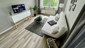 A modern living room with a white sofa, a glass coffee table, a white TV stand, and a large window with curtains. - Cozy apartment in the center (Chemnitz)