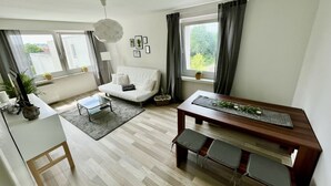 A modern living room with a white sofa, a glass coffee table, a wooden dining table with benches, and large windows with curtains. - Cozy apartment in the center (Chemnitz)