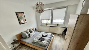 A modern bedroom with a wooden bed, grey bedding, a hanging light fixture, and a window with blinds. - Cozy apartment in the center (Chemnitz)