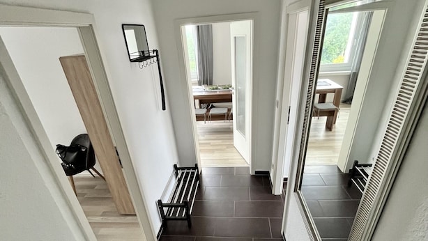 A modern hallway with dark tiled flooring, white walls, and a bench. There is a door on the left and a large mirror on the right. - Cozy apartment in the center (Chemnitz)