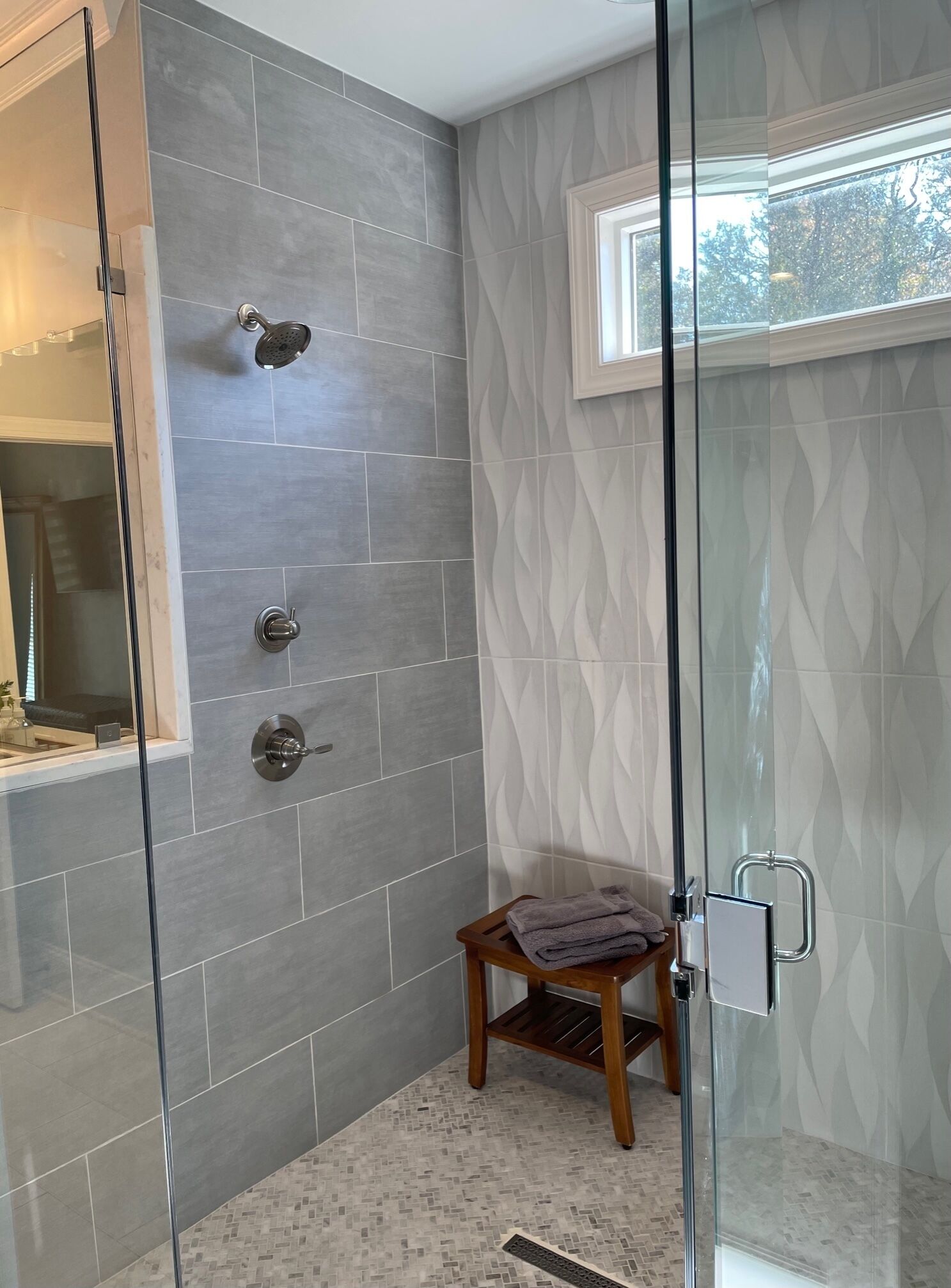 Un baño moderno con cabina de ducha de vidrio, baldosas grises, un taburete de madera pequeño y una ventana con vistas.
