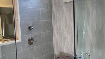 Un baño moderno con cabina de ducha de vidrio, baldosas grises, un taburete de madera pequeño y una ventana con vistas.