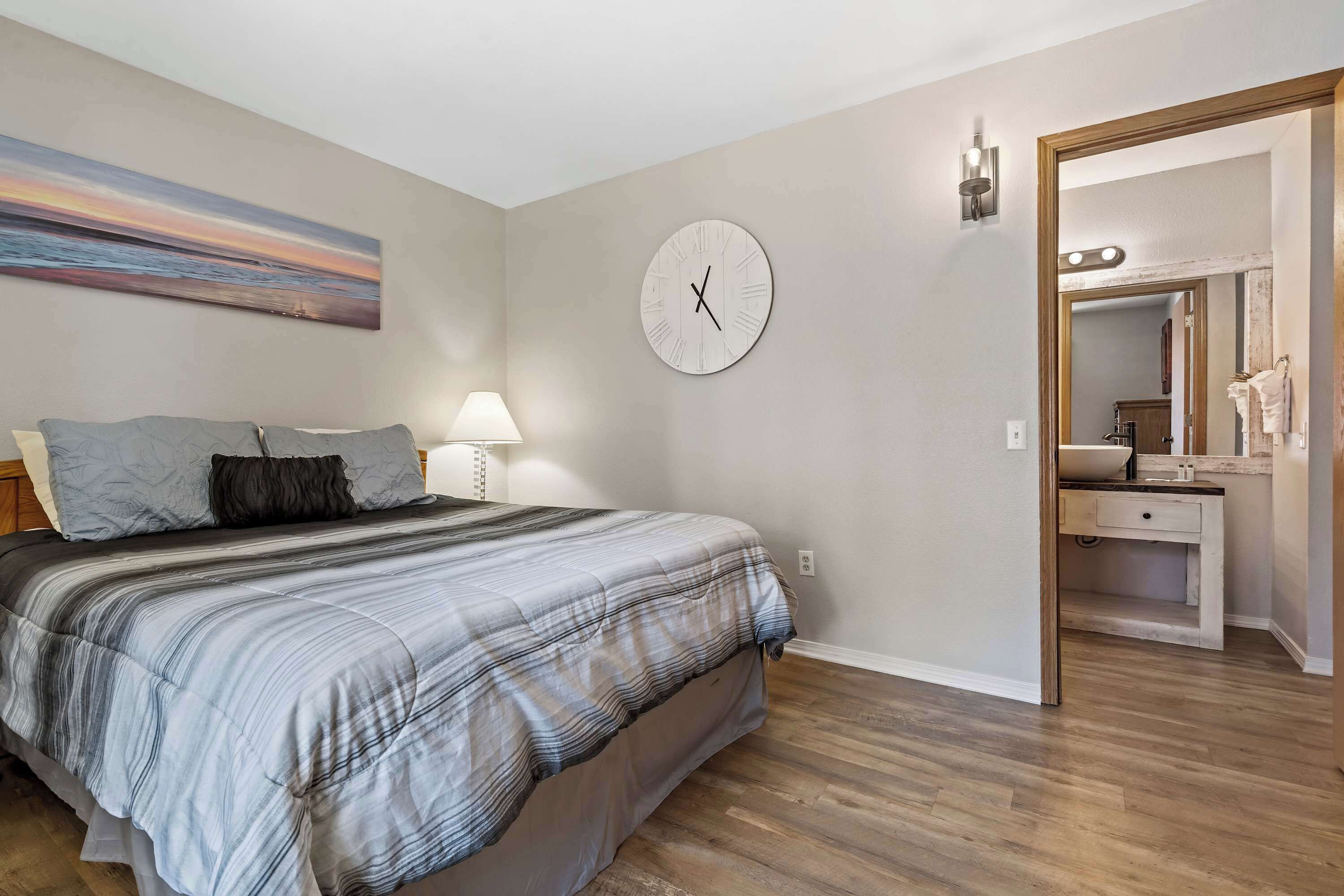 Une chambre à coucher avec un lit, une horloge murale, un tableau, et une salle de bain visible à travers une porte ouverte.