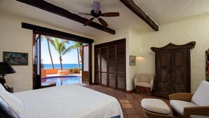 Une chambre avec un grand lit, un ventilateur de plafond, une vue sur l’océan, une piscine et une armoire en bois.