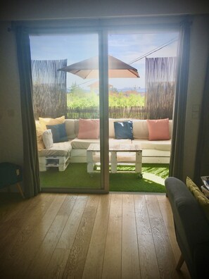 A room with a sliding glass door leading to an outdoor area with a wooden deck, a white sofa with cushions, and a small table.
