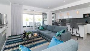 Ein modernes Wohnzimmer mit grauem Sofa, einem Couchtisch und einem gemusterten Teppich. Es gibt eine Küchenzeile mit einer Bar und einem Fernseher.