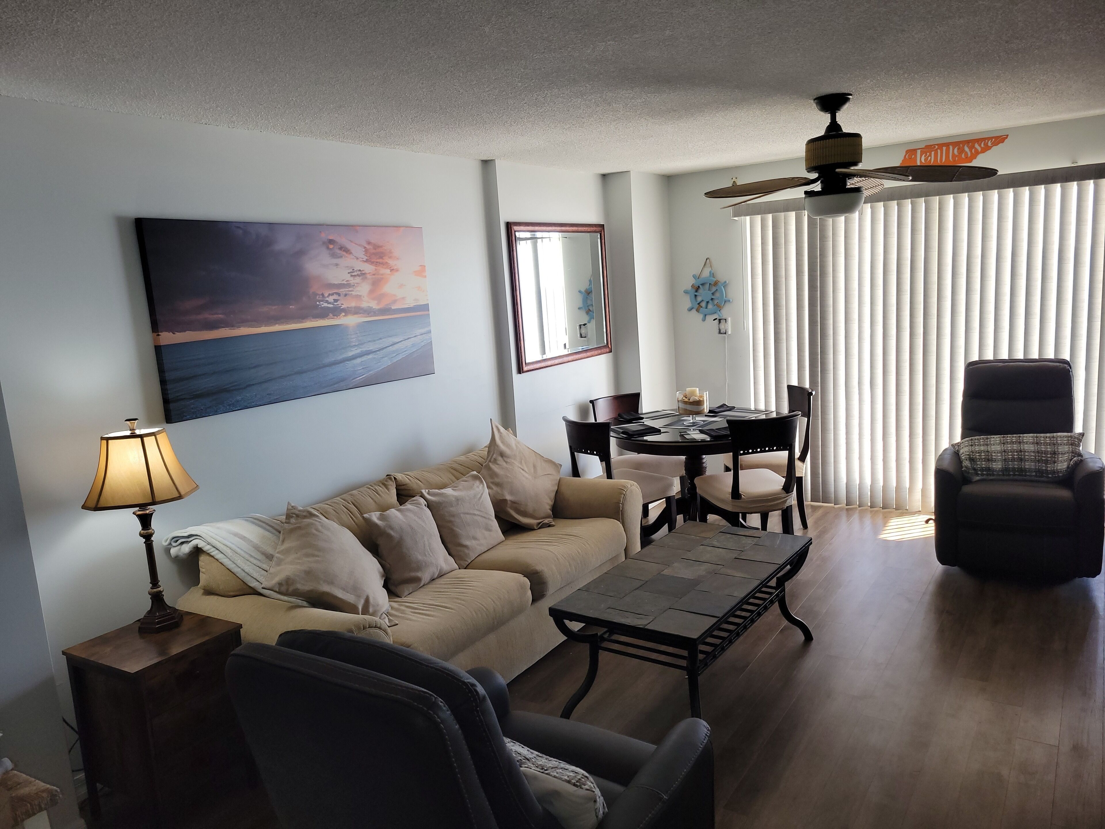 Ein Wohnzimmer mit einem beigefarbenen Sofa, einem Couchtisch, einem Esstisch mit Stühlen, einem schwarzen Sessel, einer Lampe, einem Deckenventilator, einem großen Gemälde und einem Fenster mit Jalousien.