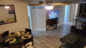 Un comedor con una mesa redonda y sillas, un televisor al fondo, y una zona de cocina con taburetes.