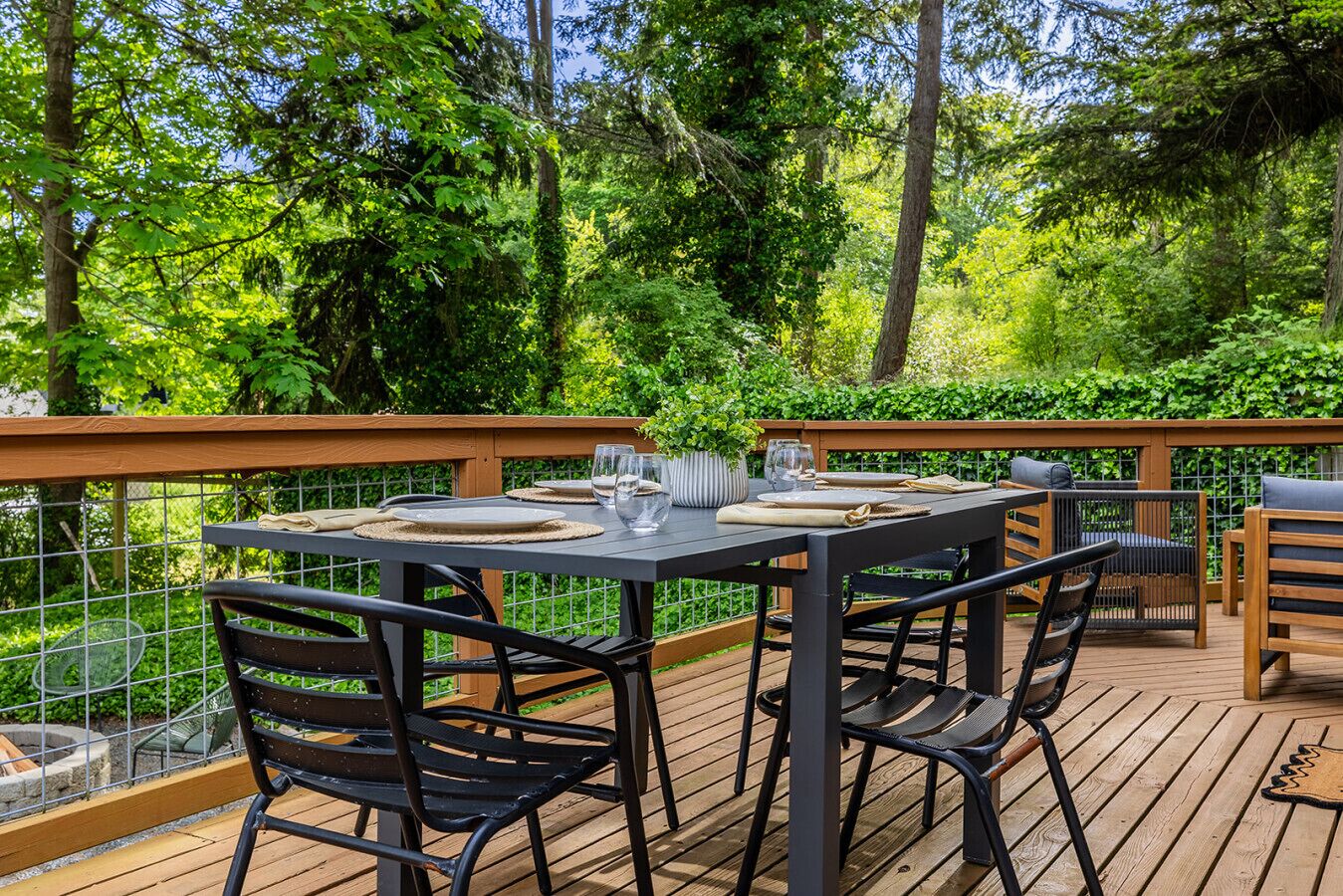 An outdoor dining area with a table set for four, surrounded by chairs, on a wooden deck with a view of lush greenery.