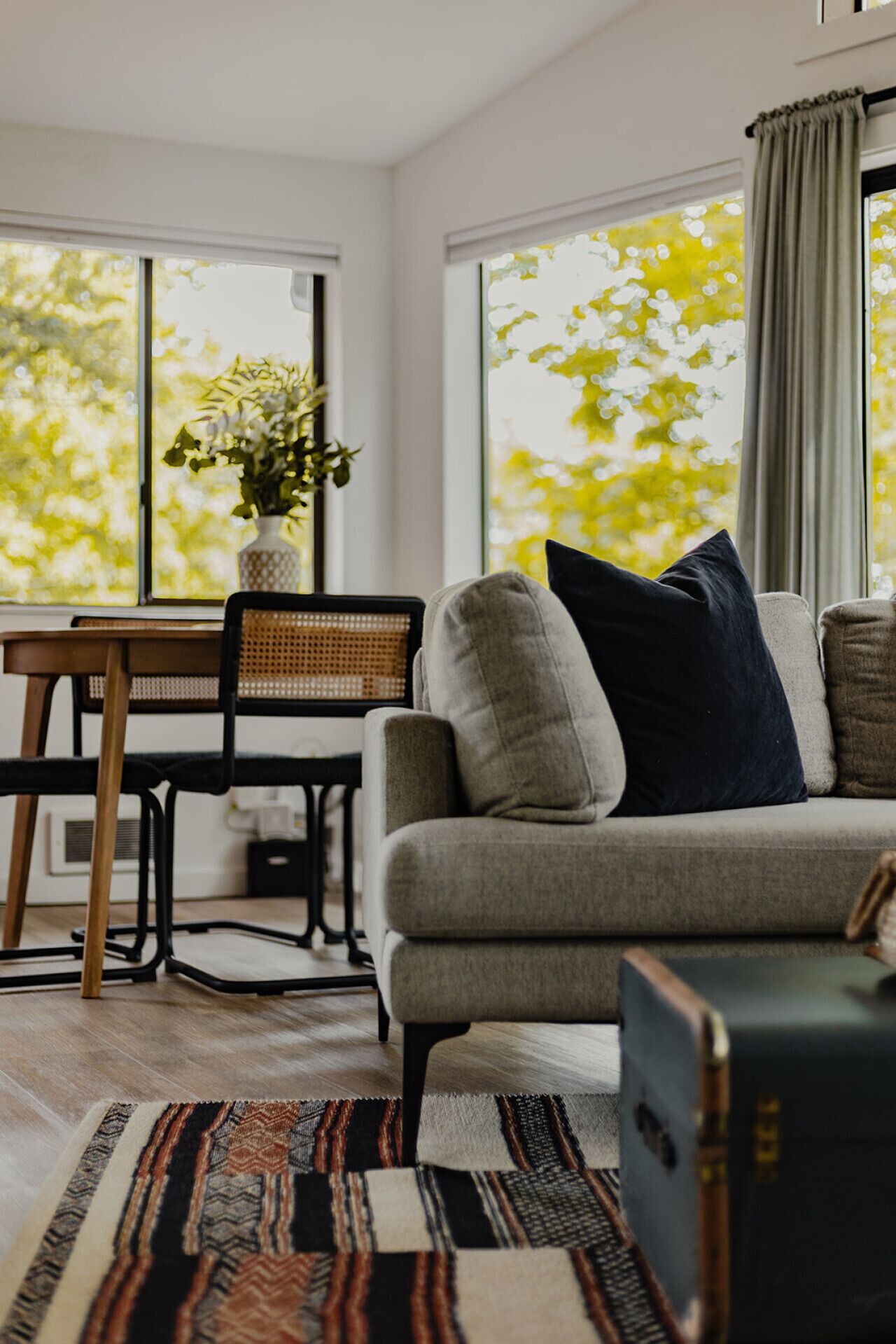 Un salon moderne avec un canapé gris, un tapis à motifs, une table à manger avec des chaises et une grande fenêtre donnant sur des arbres.