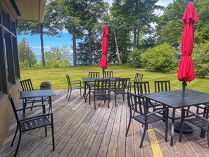 Una terraza de madera con mobiliario exterior y sombrillas rojas, con vistas a una zona ajardinada con árboles.