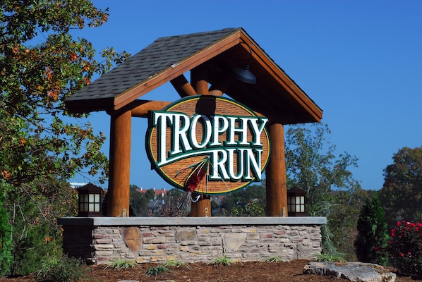 A wooden sign with the words "TROPHY RUN" in a circular design, supported by stone pillars, set against a backdrop of trees and a clear sky. - Beautiful Cabin - Secluded But Convenient (Branson)