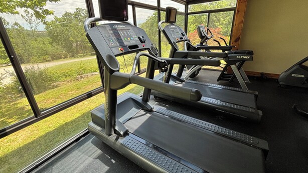 A gym with treadmills and exercise equipment, large windows with a view of greenery, and a control panel on the treadmill. - Beautiful Cabin - Secluded But Convenient (Branson)