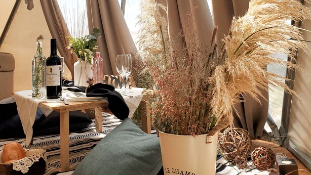 Un espacio acampado acogedor con una mesa preparada para comer, que incluye una botella de vino, copas y un jarrón decorativo con plantas secas.