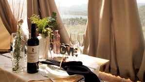 Una mesa de comedor preparada para dos, con una botella de Merlot, un florero con flores y vistas a un paisaje montañoso a través de una ventana de tienda de campaña.