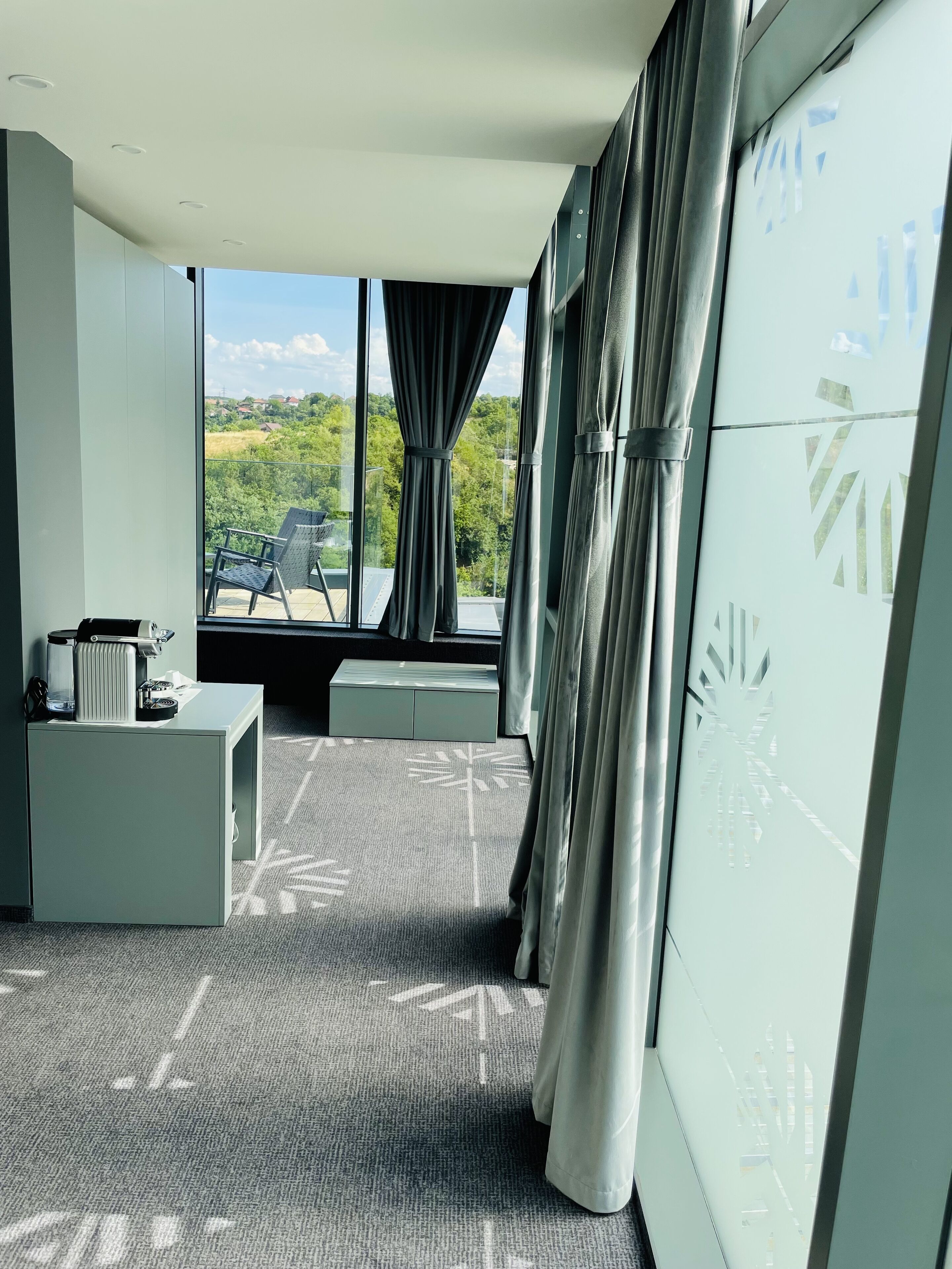Ein modernes Hotelzimmer mit großem Fenster, einer Sitzecke mit Sofa und Sesseln, einem kleinen Tisch mit Wasserkocher und einem gemusterten Teppich.