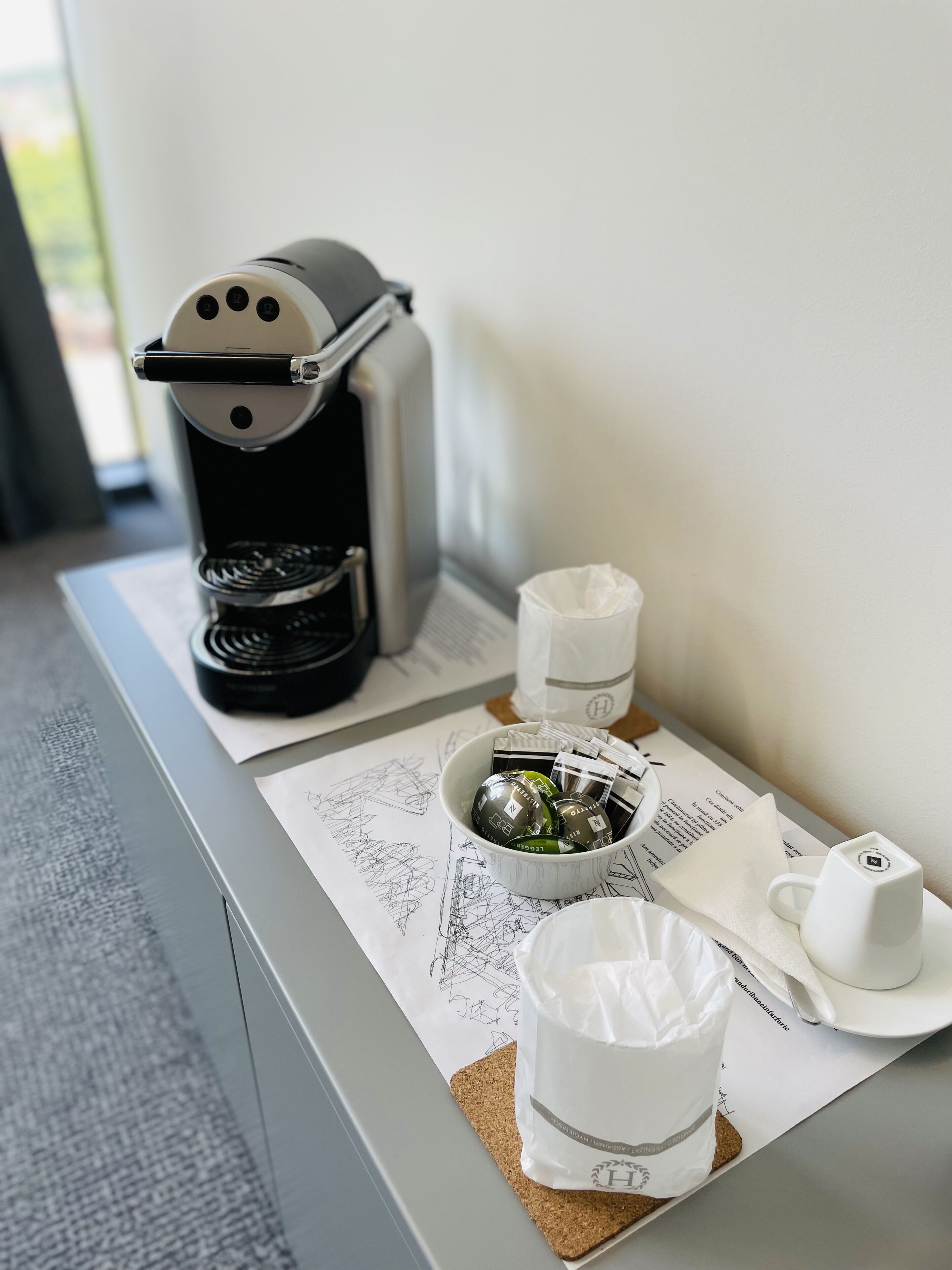 A coffee maker, a cup, a mug, and a small bowl with coffee pods on a table with a menu.