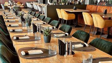 A restaurant with long wooden tables, green upholstered chairs, and hanging plants.