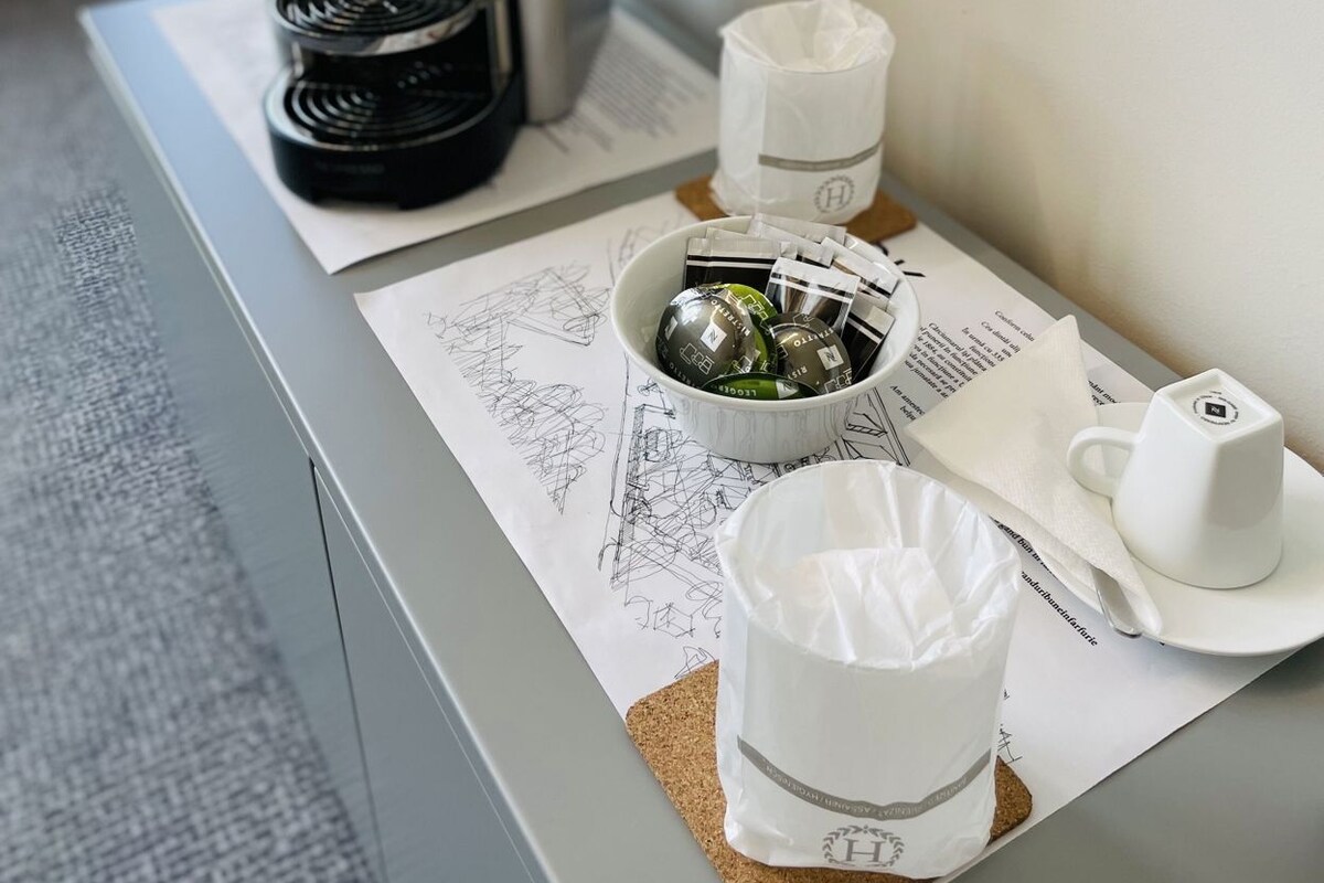 Una cafetera, una taza, un vaso y una bolsa de papel sobre una mesa con una ventana al fondo.