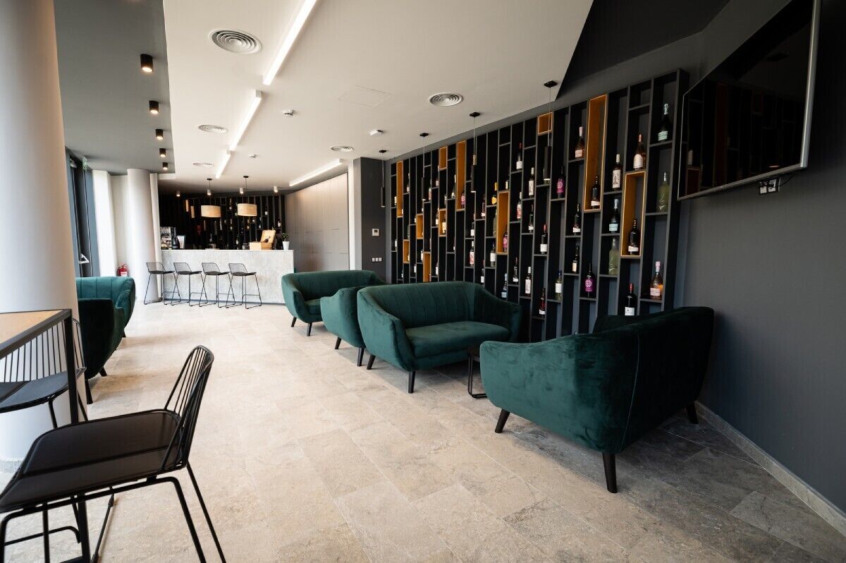 A modern interior with a reception area, green upholstered chairs, a bar, and a wall of wine bottles.