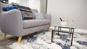 Un salon moderne avec un canapé gris, des coussins à motifs, une table basse en verre et un tapis aux motifs bleus et blancs.