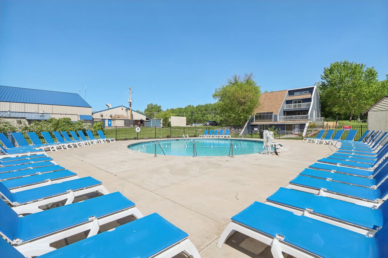 A pool area with blue lounge chairs arranged around it, a clear sky, and buildings in the background.