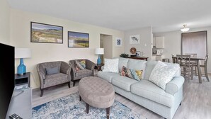 A living room with a sofa, two armchairs, a coffee table, and a TV. There are framed pictures on the wall, a dining area with a table and chairs, and a kitchen area in the background.