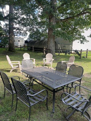 A metal table with chairs in a grassy outdoor area, surrounded by trees and a swing set.