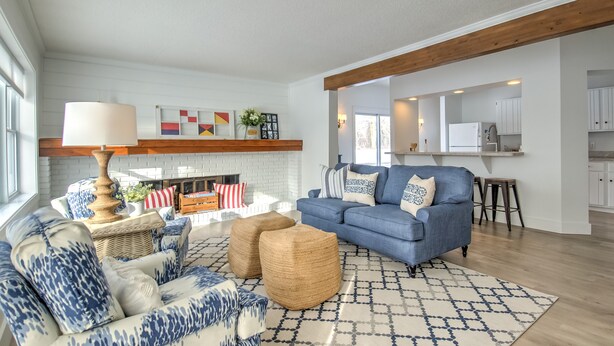 A spacious living room with a blue sofa, a coffee table, and a fireplace. There is a kitchen area with white cabinets and a wooden ceiling beam.