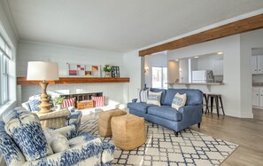 Amplia sala de estar con sofá azul, una mesa de centro y una chimenea. Se observa una zona de cocina con armarios blancos y una viga de techo de madera.