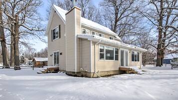 A single-story house with a gabled roof and a chimney, surrounded by snow-covered ground and leafless trees.