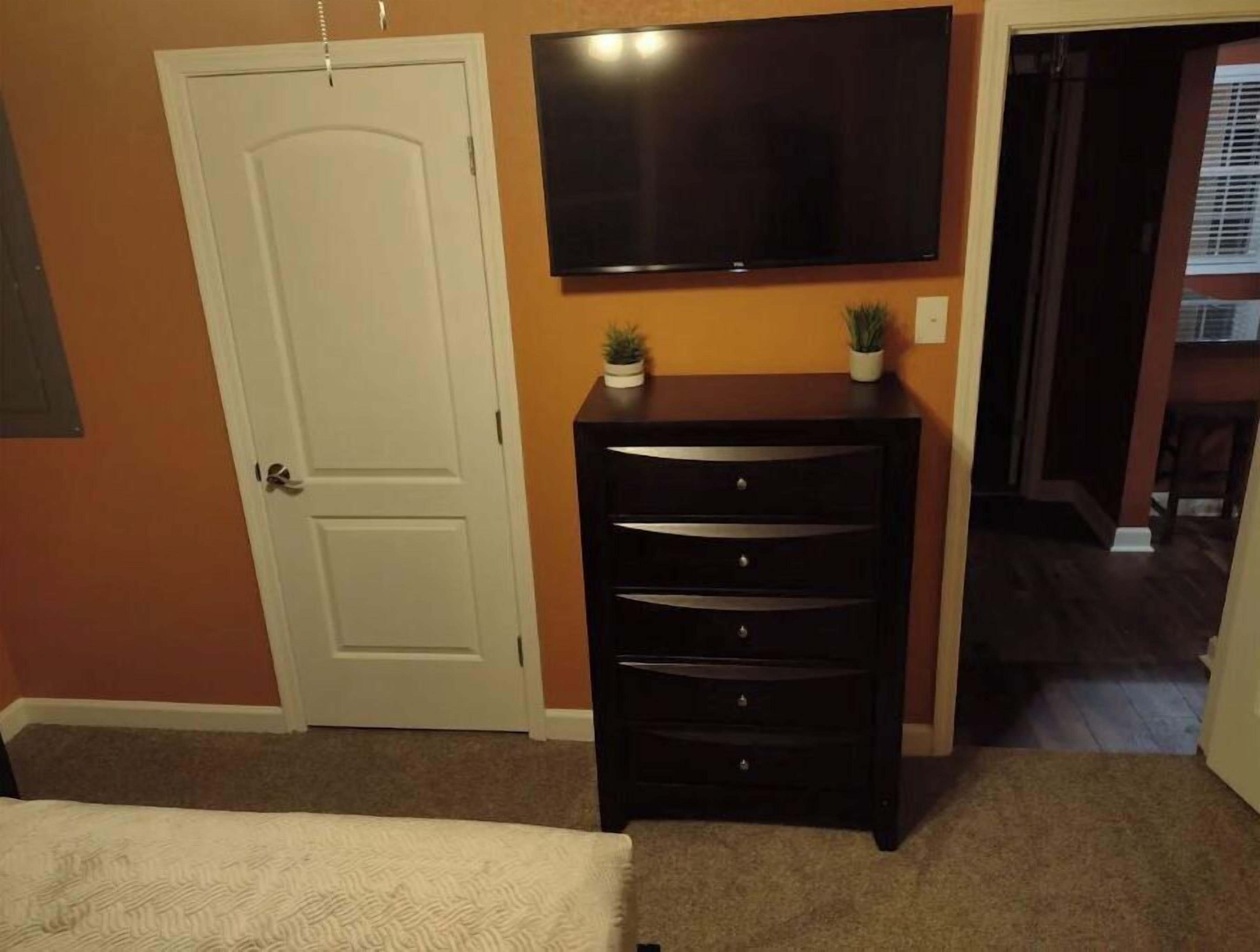 Un dormitorio con una cama, un aparador, un televisor montado en la pared y una puerta.