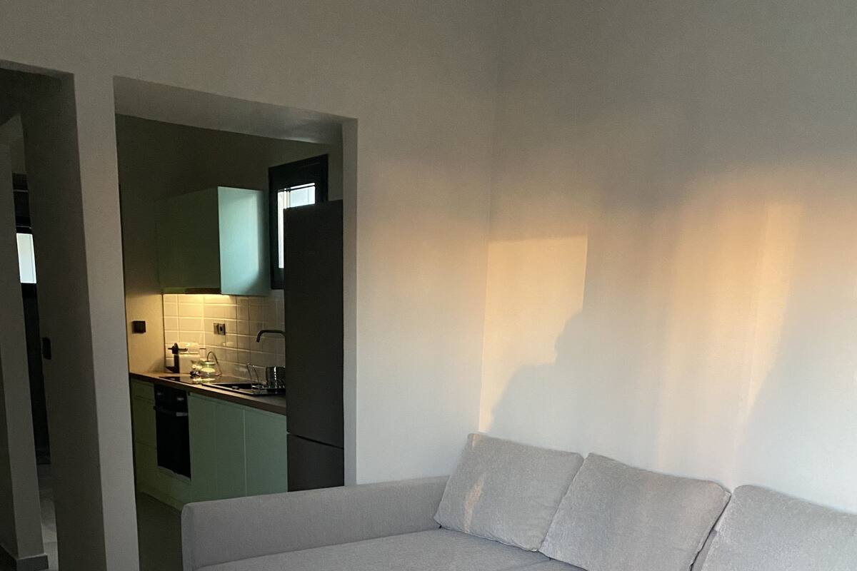 Un salon moderne avec un canapé d’angle gris, une table basse en bois et une cuisine visible au fond.
