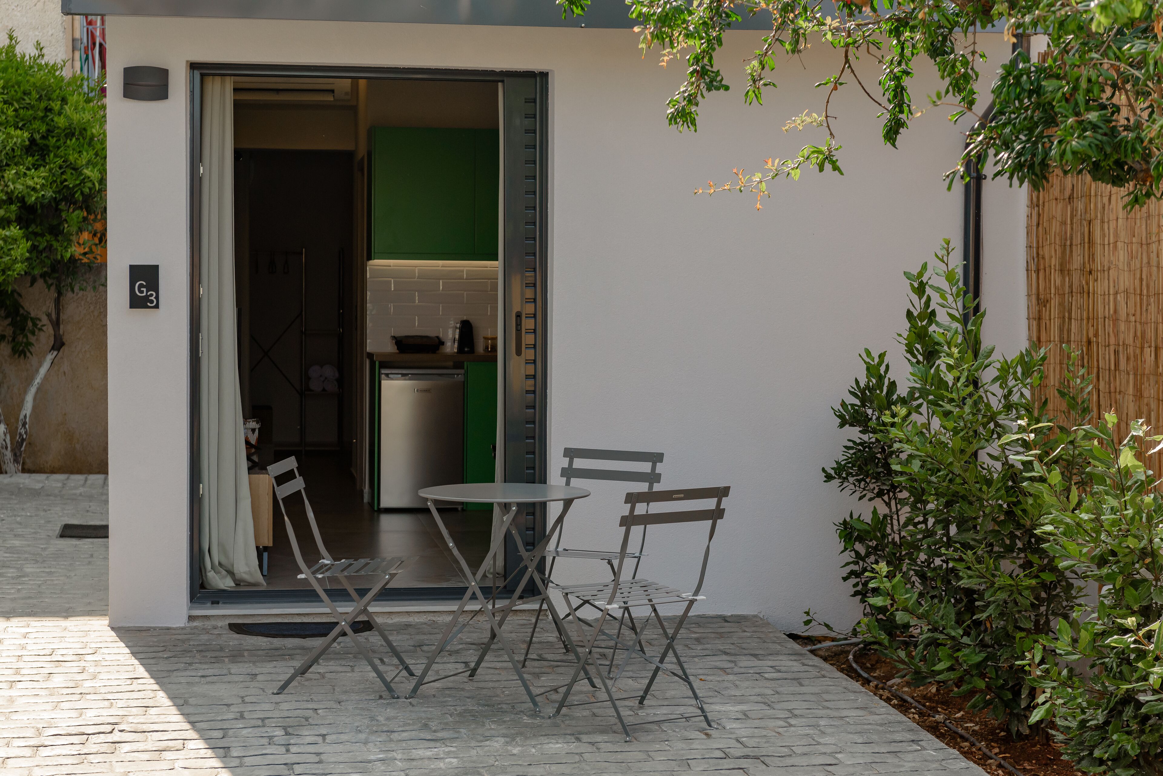 Un pequeño espacio al aire libre con dos sillas y una mesa, una puerta que da a una cocina, y una valla de bambú.