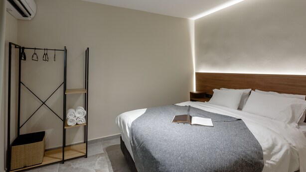 A modern hotel room with a neatly made bed, a wooden headboard, a built-in shelf with towels, and a wall-mounted air conditioner. - Galio Suites Airport by Airstay (Spata-Artemida)