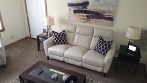 Ein Wohnzimmer mit einem beigefarbenen Sofa, zwei gemusterten Zierkissen, einem Glastisch mit Fernbedienung, einer Lampe und einem Gemälde an der Wand.