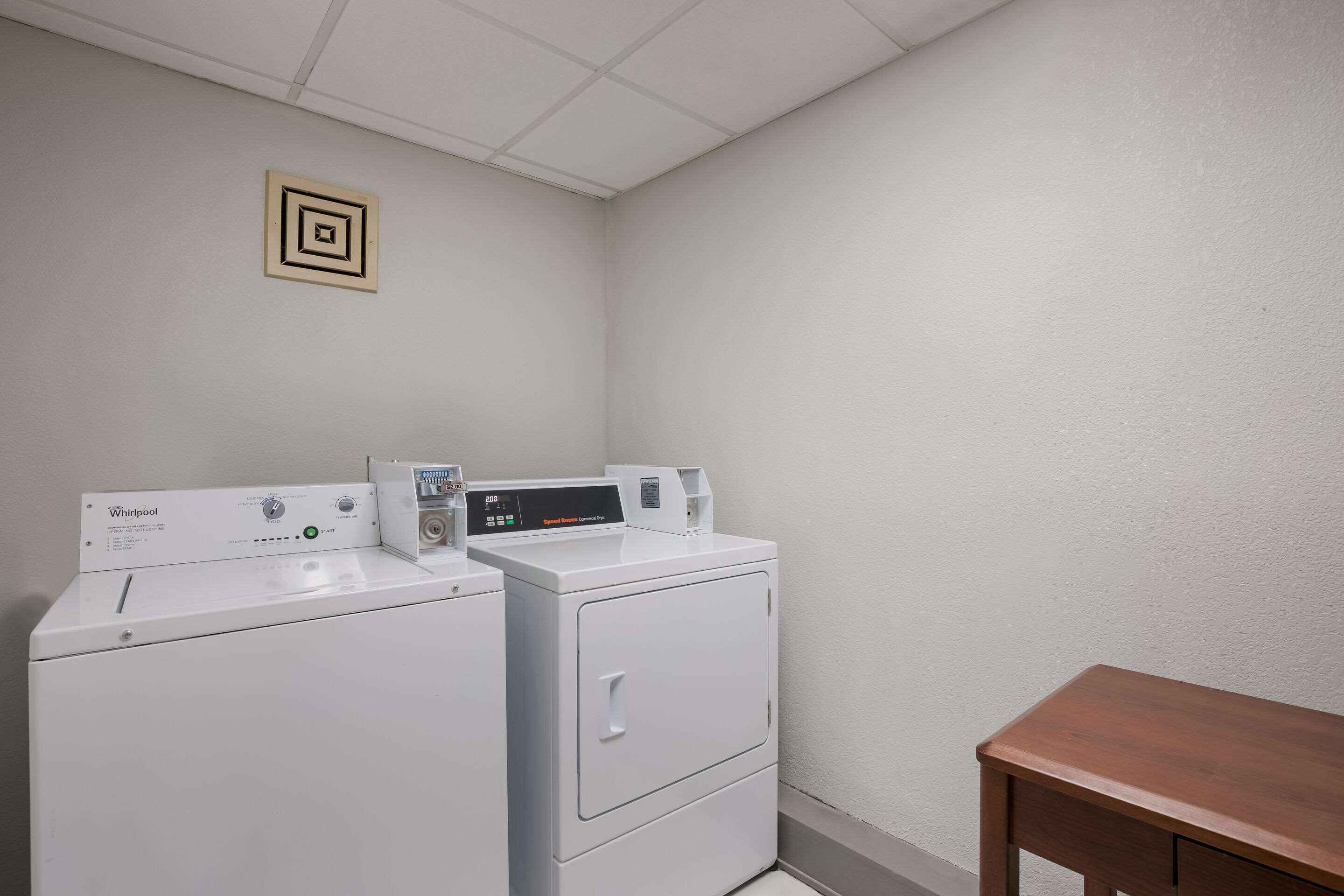A laundry room with two Whirlpool washers and dryers, a wooden bench, and a small decorative item on the wall.
