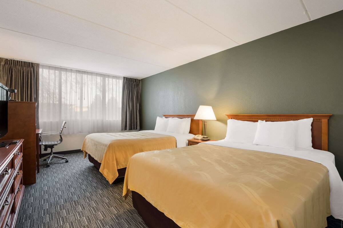 A hotel room with two beds, a desk, a chair, and a lamp.