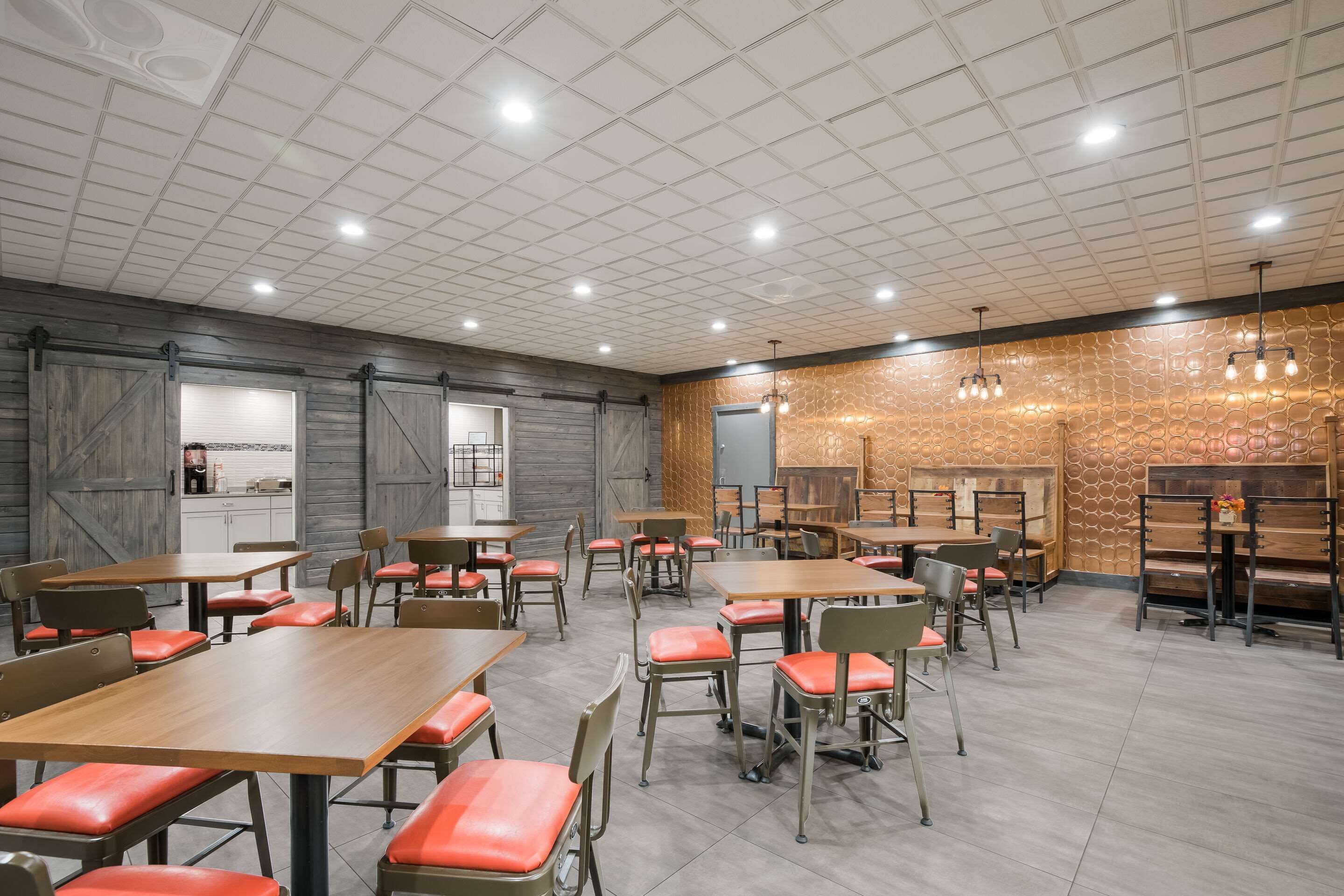 A spacious dining area with wooden tables and red cushioned chairs, a kitchen area, and a decorative wall with a geometric pattern.