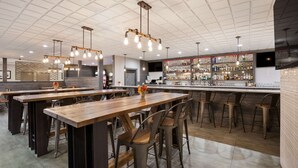 Un área de bar moderna con varias mesas de madera, taburetes de bar metálicos, una barra bien surtida y lámparas colgantes.