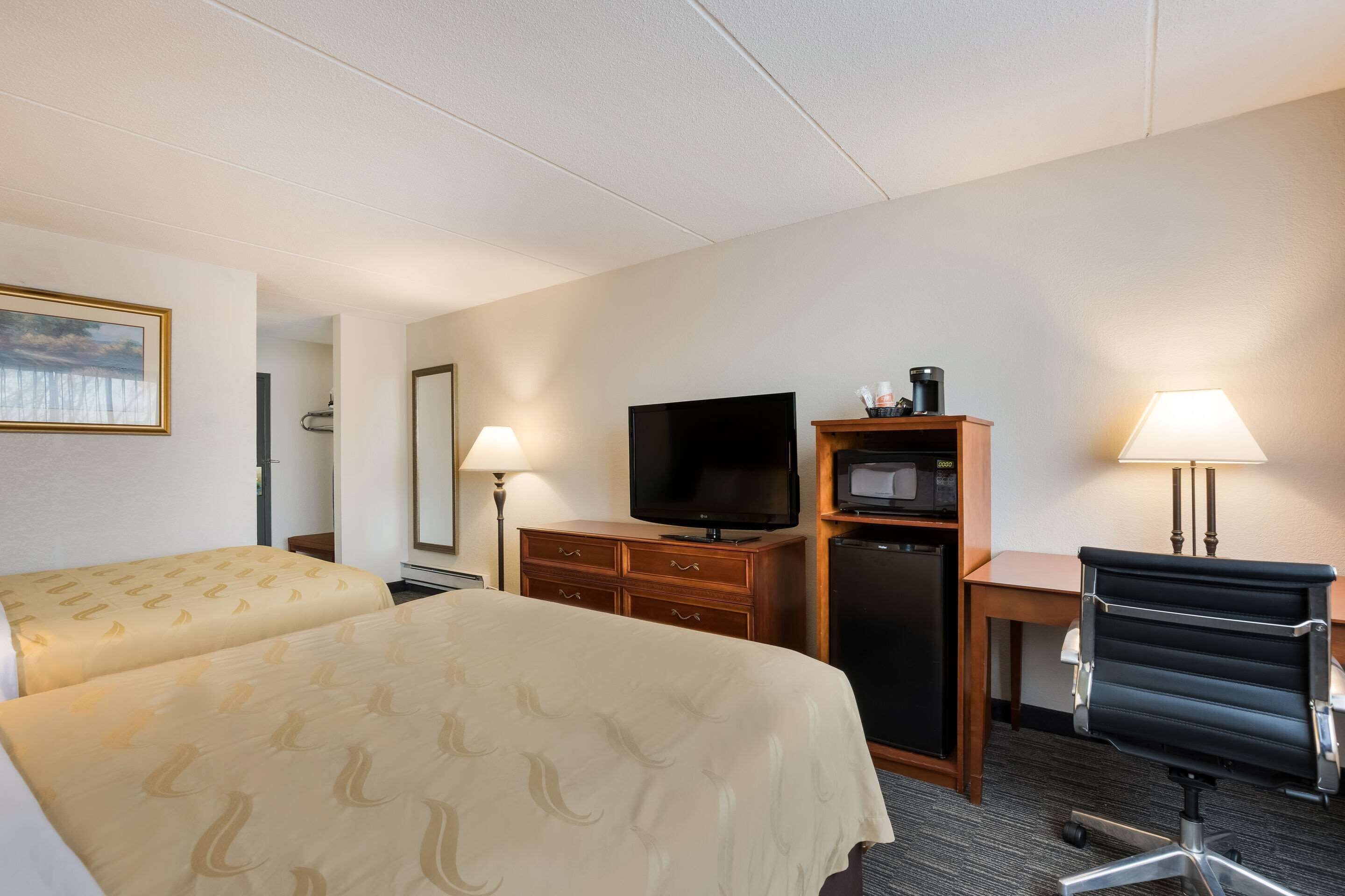Habitación de hotel con dos camas, un escritorio con computadora, televisor, refrigerador y dos lámparas.