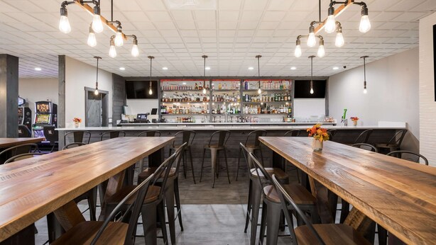 Una zona de bar con mesas y sillas de madera, una barra con varias botellas y lámparas colgantes.