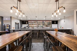 A bar area with wooden tables and chairs, a bar counter with various bottles, and hanging light fixtures.