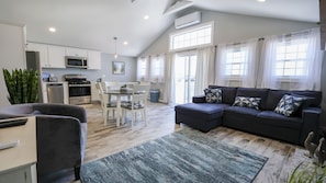 A modern living room with a dark sofa, a dining table, and a kitchen area with white cabinets and appliances.