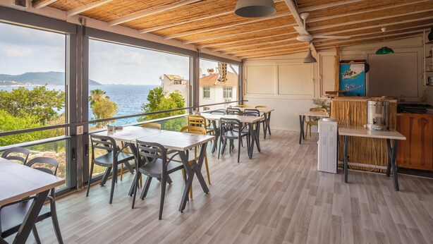 A modern dining area with wooden flooring, large windows offering a view of the sea, and a kitchen area with a refrigerator and a blue sign. - Nest Gunay Otel (Kas)