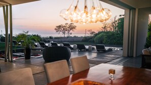Un espace de restauration en plein air offrant une vue sur l’océan au coucher du soleil, avec une table en bois, des chaises et un luminaire suspendu.
